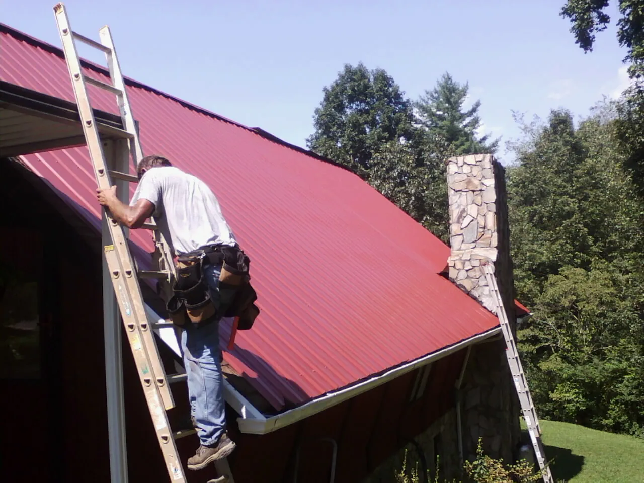 Roofer on a ladder beside a red metal roof and stone chimney — Soffit & Fascia Repair by Roofing Brothers in Sweeny
